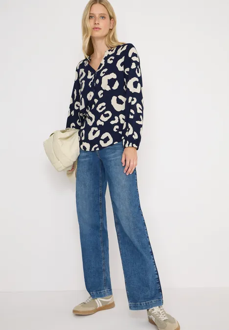 Frau mit gemusterter Bluse, blauer Jeans und Turnschuhen, Beutel in der Hand.